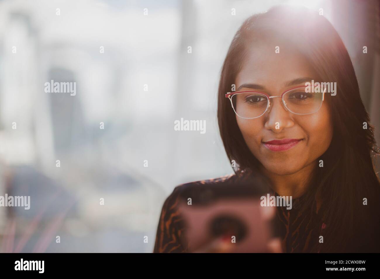 Femme d'affaires souriante utilisant un smartphone à la fenêtre ensoleillée Banque D'Images