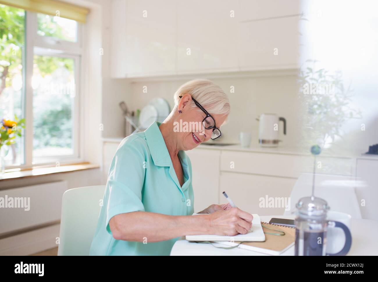 Femme âgée écrivant dans le journal à la table de cuisine du matin Banque D'Images