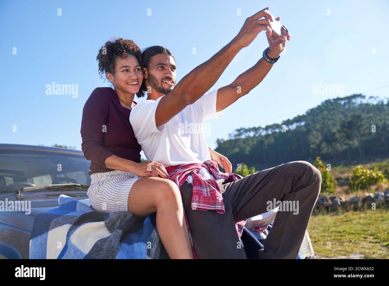 Un jeune couple heureux prend le selfie sur le capot de la voiture à route ensoleillée Banque D'Images