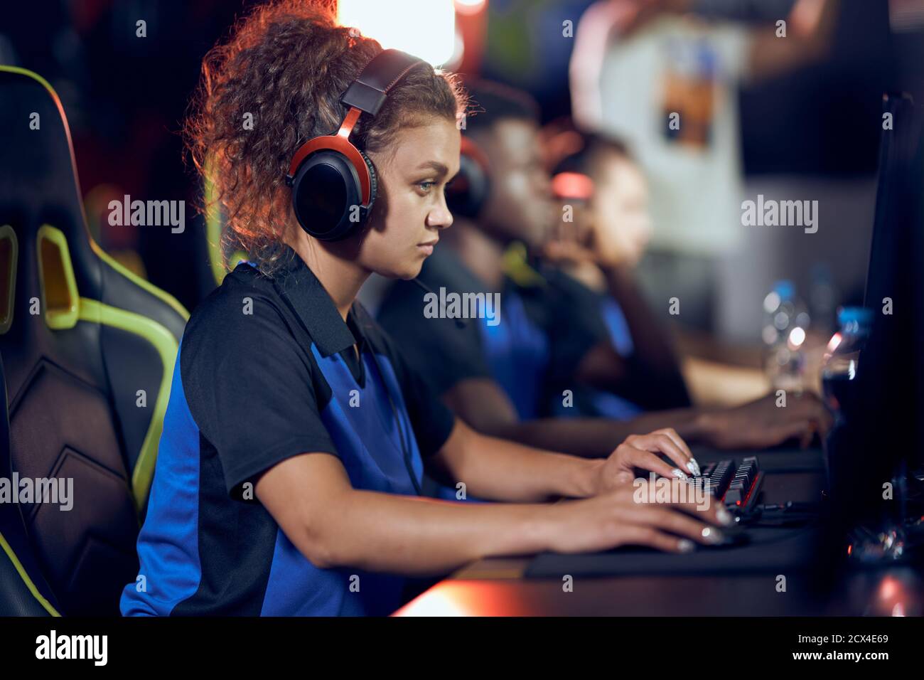 Vue latérale d'une jeune fille de course mixte ciblée, d'une joueuse de cybersport portant un casque jouant à des jeux vidéo en ligne, participant à un tournoi eSport tout en étant assis dans un club de jeu ou un café Internet Banque D'Images