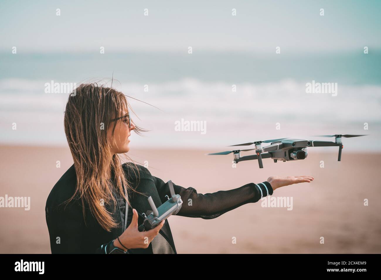 Jeune femme moderne pilote dans la veste noire pays drone sur sa main avec télécommande près de l'océan. Passe-temps en plein air. Banque D'Images