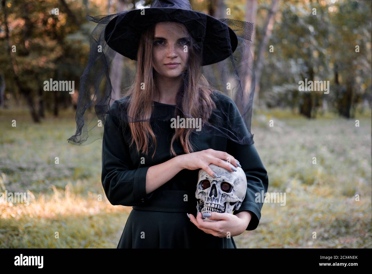 Jeune belle femme mignonne en robe sombre et chapeau de sorcière tient un crâne dans ses mains. Costume de fête d'Halloween. Forêt, parc avec arbres d'automne Banque D'Images