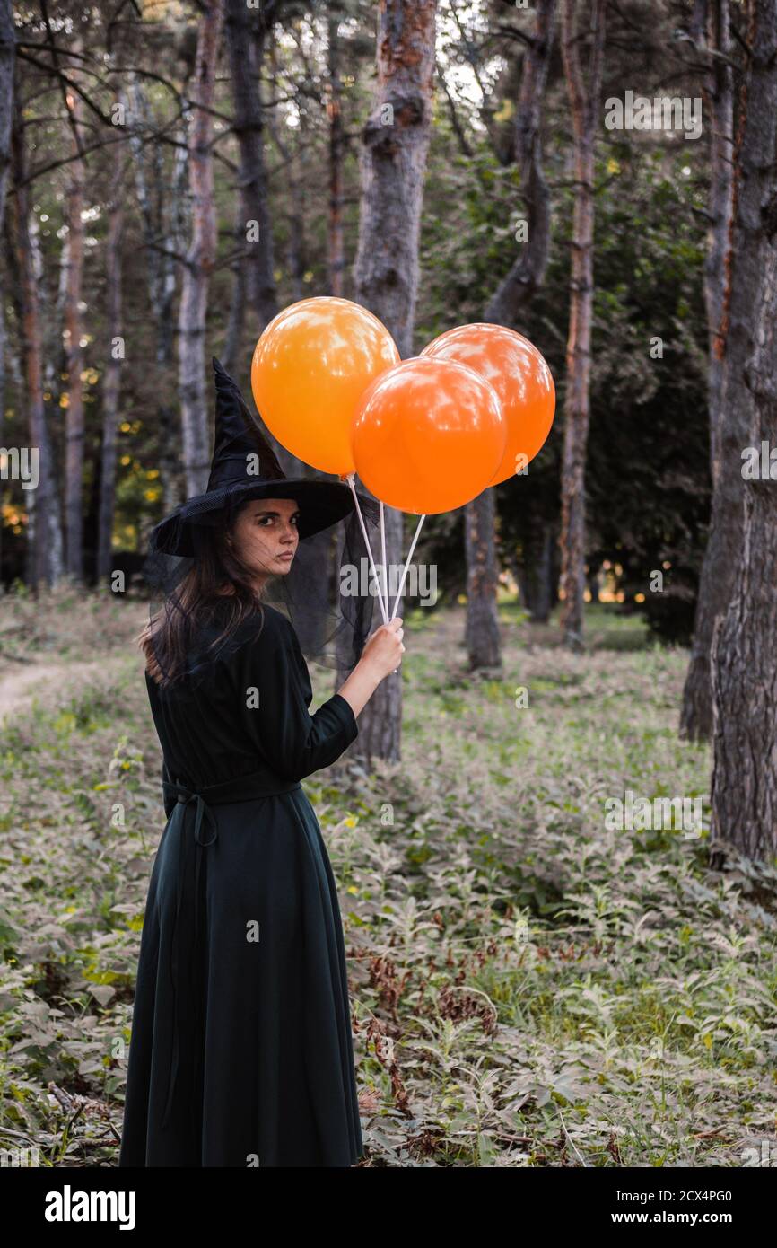 Jeune mignonne belle femme en robe sombre et chapeau de sorcière tient des ballons d'orange dans ses mains. Costume de fête d'Halloween. Forêt, parc avec arbres d'automne Banque D'Images