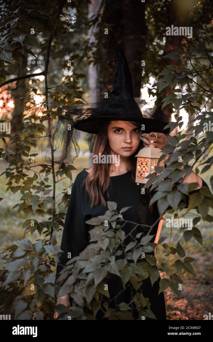Une jeune femme en robe sombre et chapeau de sorcière tient une lanterne avec des bougies dans ses mains et illumine la forêt. Costume de fête d'Halloween. Garez-vous avec l'autom Banque D'Images