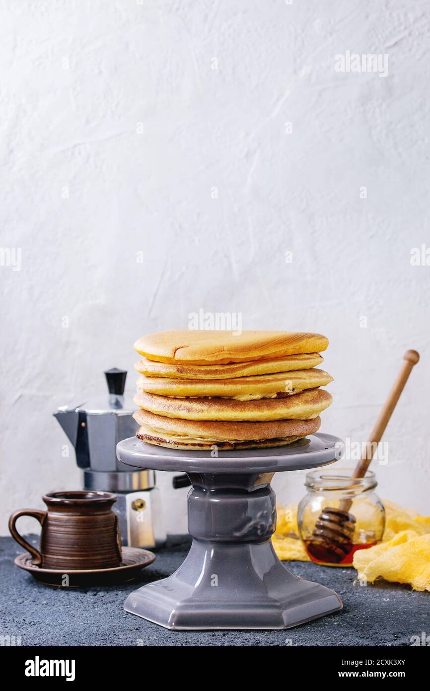 Pile de crêpes américaines maison au curcuma jaune ombré servies sur un support à gâteau avec une cafetière, un pot de miel et un pot en bois sur du béton gris textu Banque D'Images