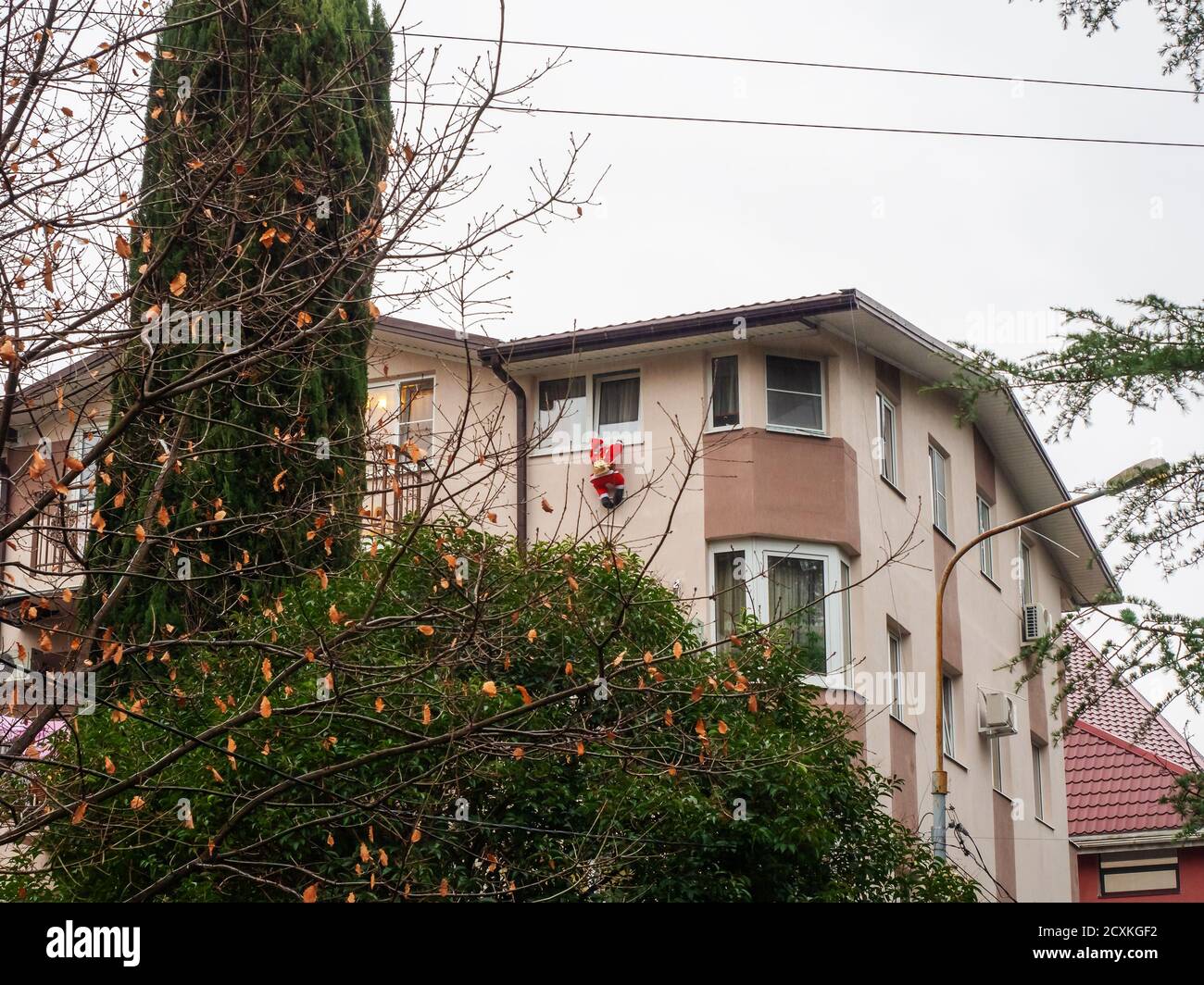 Une maison beige avec le Père Noël suspendu d'une fenêtre se dresse parmi les arbres verts et avec des feuilles mortes. Banque D'Images