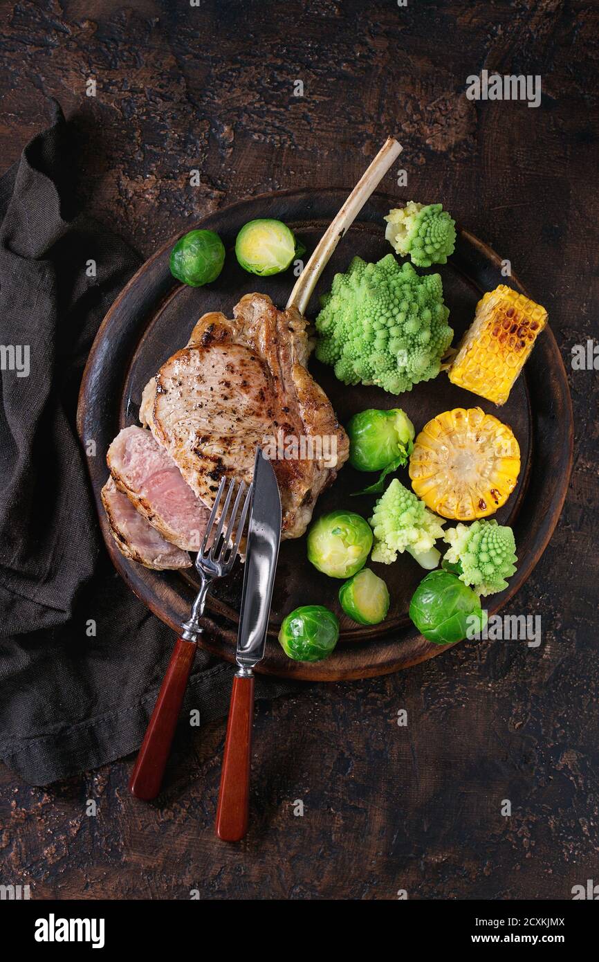 Tranches de veau grillé Steak tomahawk avec légumes choux de Bruxelles, romanesco et épis de maïs servi avec couverts en bois servant à découper sur plus de Banque D'Images