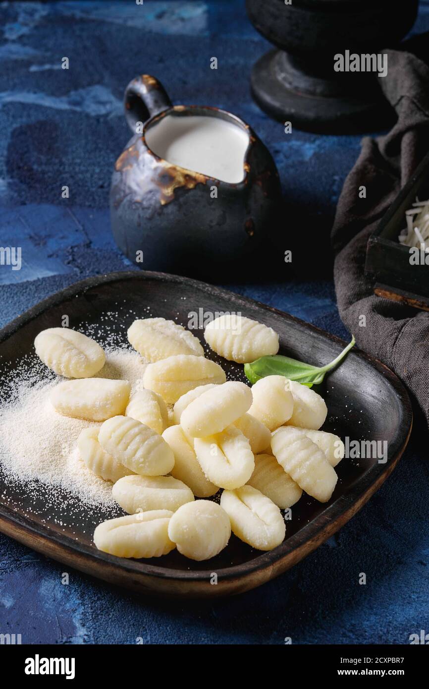 Gnocchi de pomme de terre cru non cuit dans des assiettes en bois noir avec ingrédients. Farine, parmesan râpé, basilic, carafe de crème sur fond de béton bleu foncé Banque D'Images