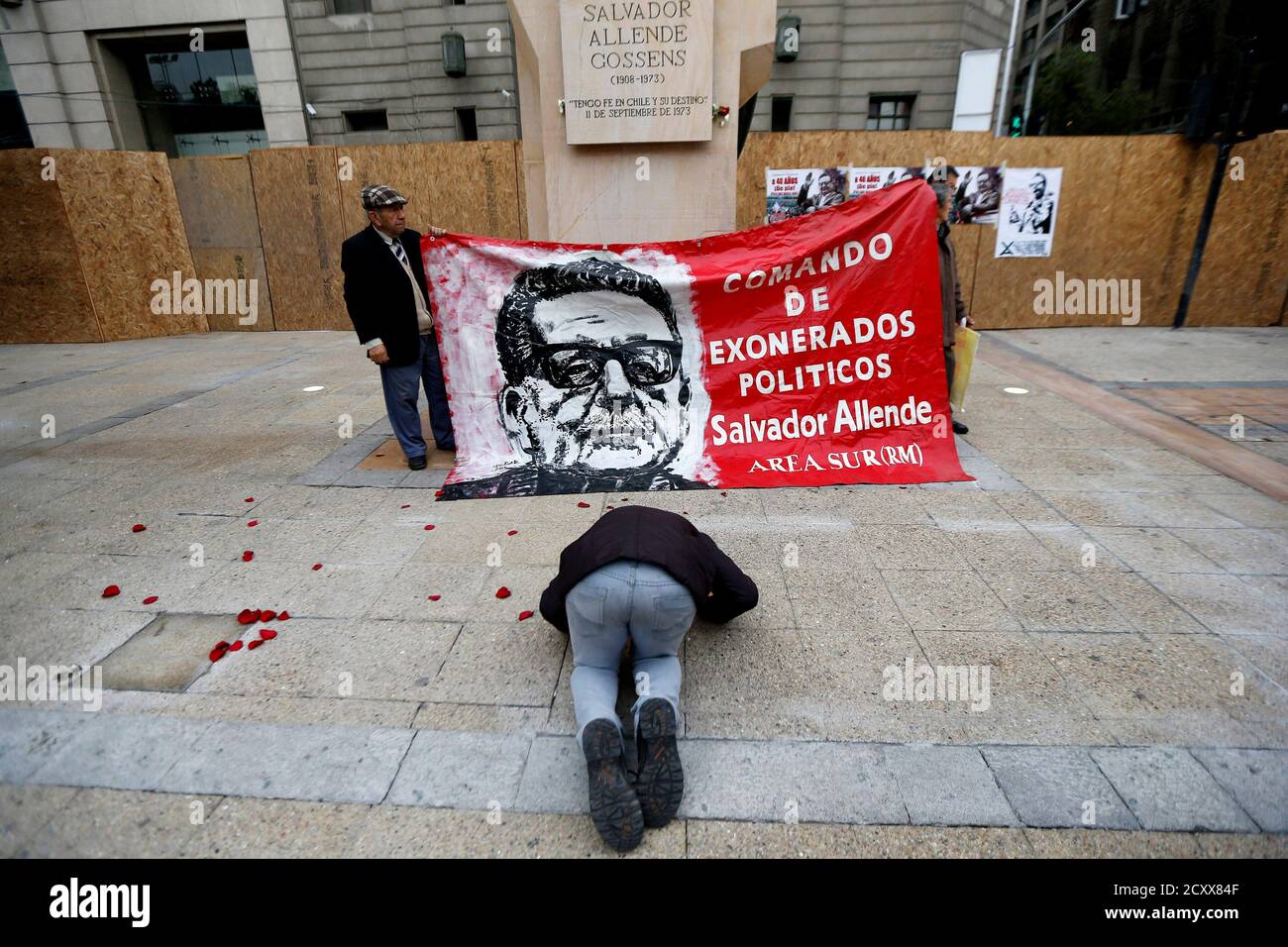 A man kneels as people hold up a banner bearing a portrait of 