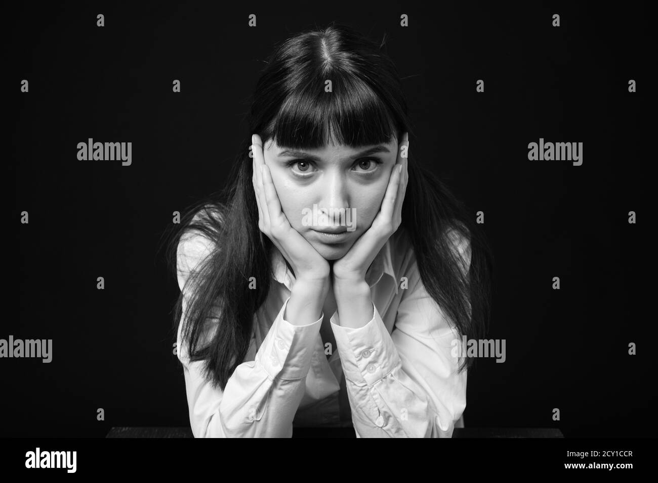 Portrait studio d'une jolie femme brune dans une chemise blanche, assise à la table, sur un fond noir Uni, regardant sérieusement l'appareil photo Banque D'Images