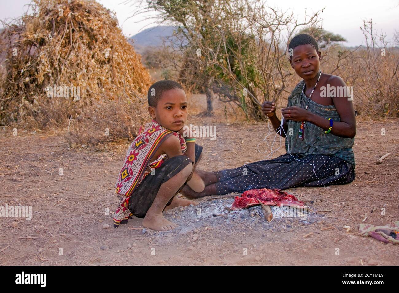 Une femme qui cuisine de la viande sur un feu de camp à côté d'elle est assise un petit enfant, le Hadza, ou Hadzabe - sont un groupe ethnique indigène dans le centre-nord de la Tanzanie Banque D'Images