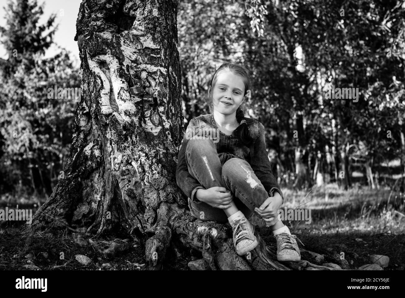 Petite fille assise sur les racines d'un bouleau. Photo en noir et blanc. Banque D'Images