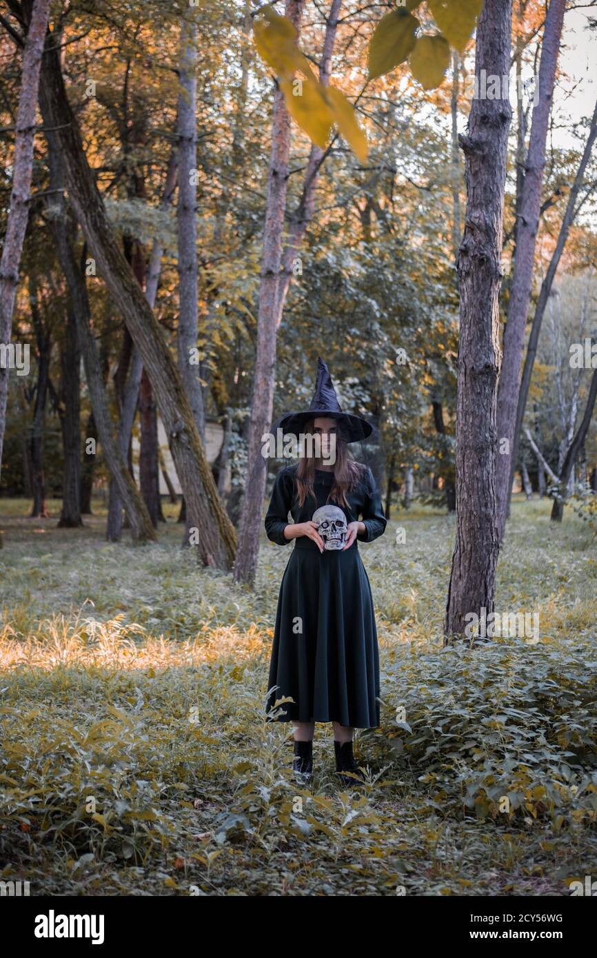 Jeune belle femme mignonne en robe sombre et chapeau de sorcière tient un crâne dans ses mains. Costume de fête d'Halloween. Forêt, parc avec arbres d'automne Banque D'Images