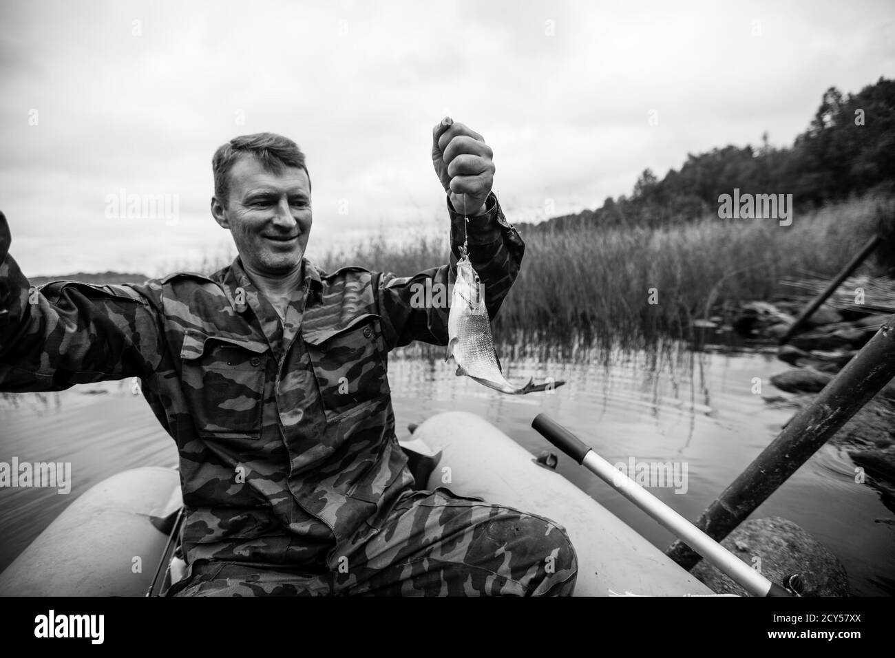 Homme dans la canne de pêche de camouflage sur la rivière sur un bateau en caoutchouc. Photographie en noir et blanc. Banque D'Images