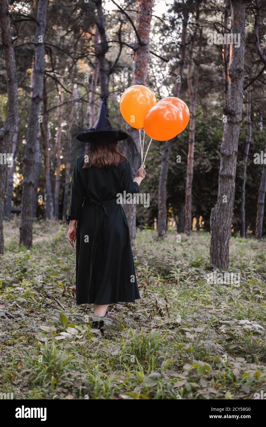 Jeune mignonne belle femme en robe sombre et chapeau de sorcière tient des ballons d'orange dans ses mains. Costume de fête d'Halloween. Forêt, parc avec arbres d'automne Banque D'Images