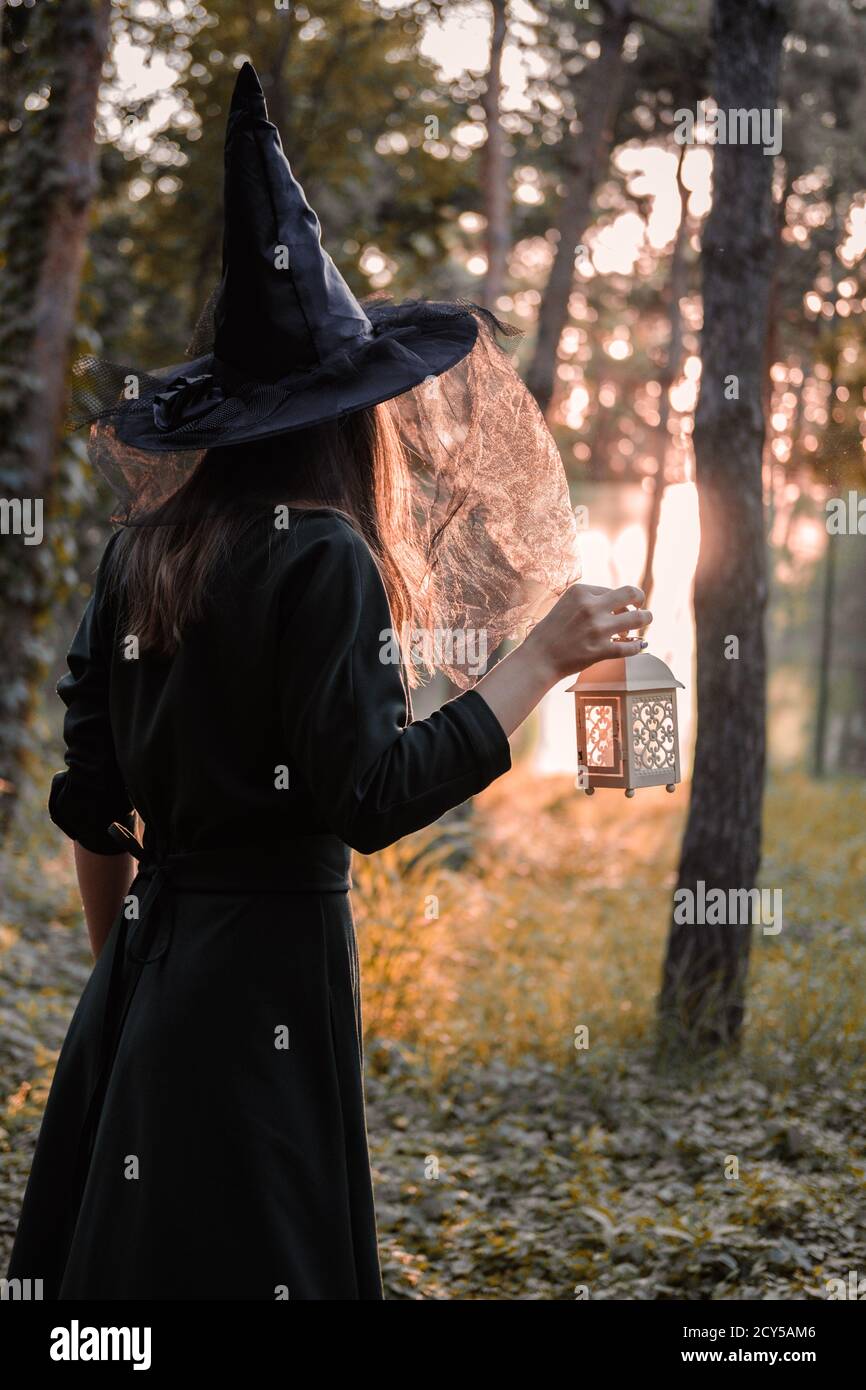 Une jeune femme en robe sombre et chapeau de sorcière tient une lanterne avec des bougies dans ses mains et illumine la forêt. Costume de fête d'Halloween. Garez-vous avec l'autom Banque D'Images