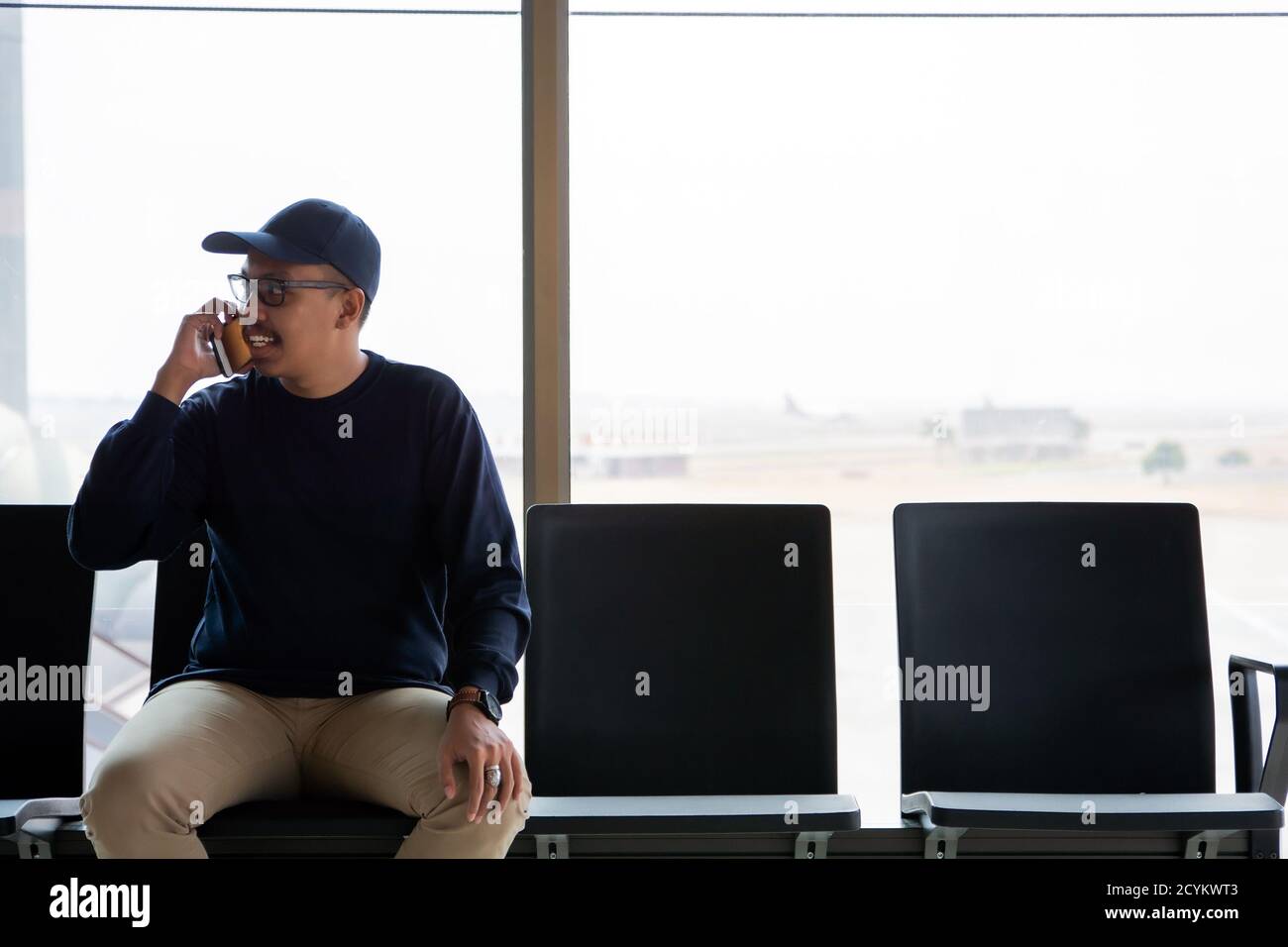 Un jeune homme asiatique joyeux attend l'avion à l'aéroport. Il est assis sur le banc et souriant. Guy utilise un téléphone intelligent pour prendre un appel téléphonique Banque D'Images