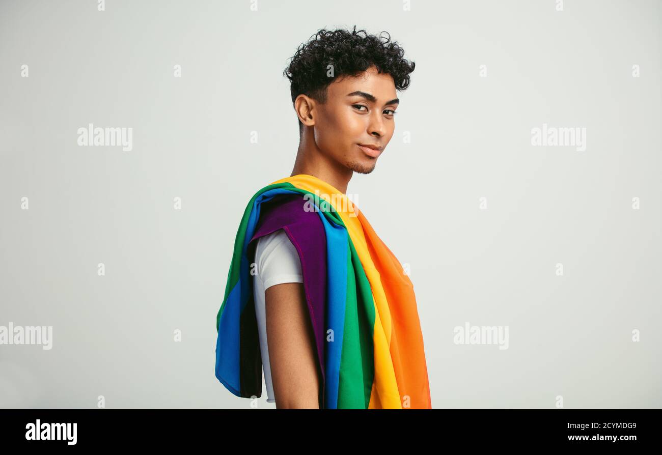 Beau jeune homme avec fierté mouvement LGBT Rainbow drapeau sur l'épaule contre fond blanc. Homme avec un drapeau de fierté gay regardant la caméra. Banque D'Images