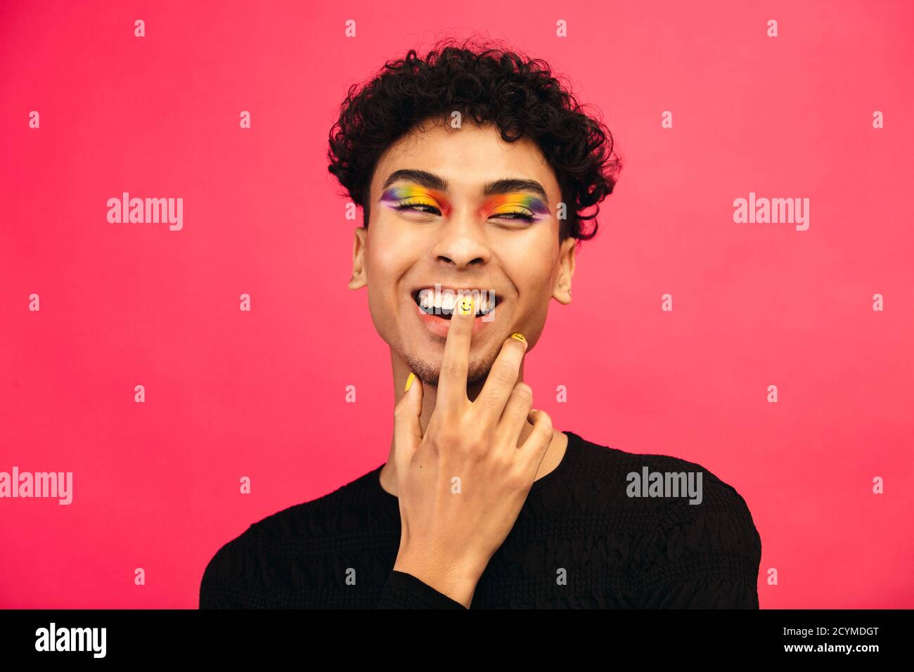 Jeune homme heureux portant une ombre à l'œil et une peinture pour ongles de visage de sourire de couleur arc-en-ciel. Homme gay souriant avec main sur le visage sur fond rouge. Banque D'Images