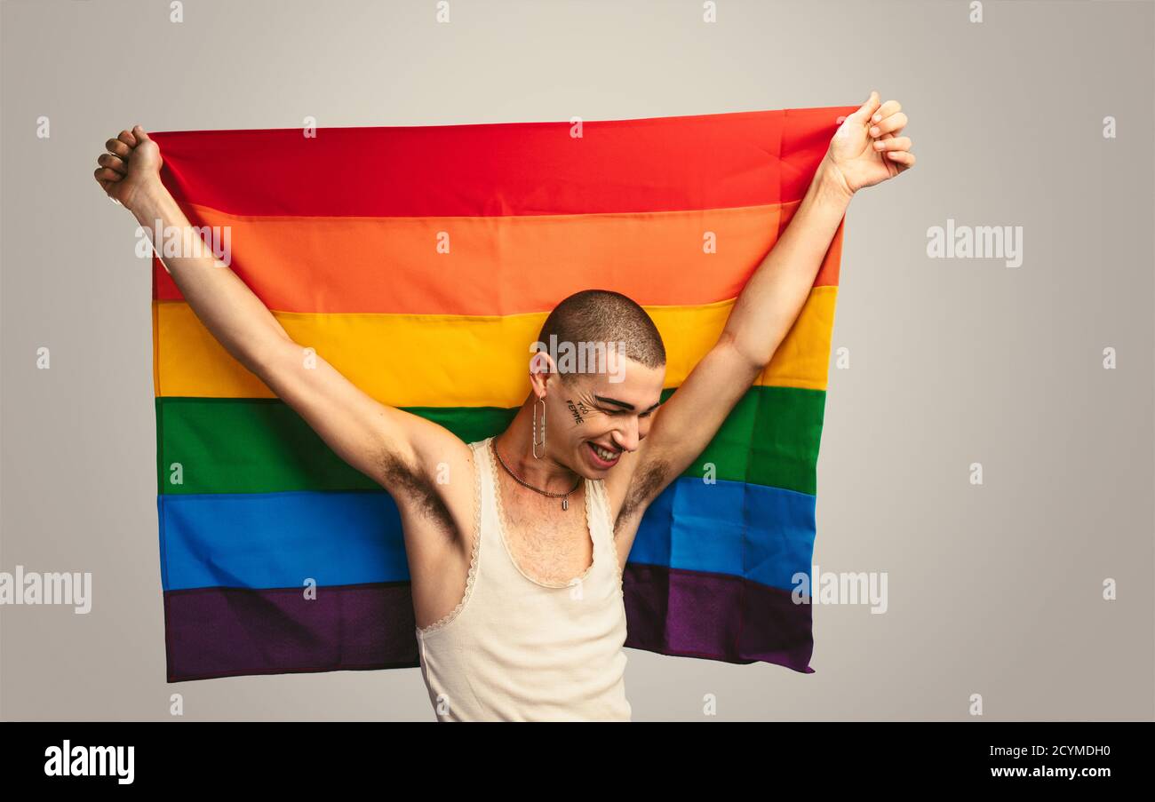 Jeune homme avec débardeur et boucle d'oreille portant un drapeau de fierté, regardant vers le bas et souriant. Homme gay souriant avec un drapeau lgbt. Banque D'Images