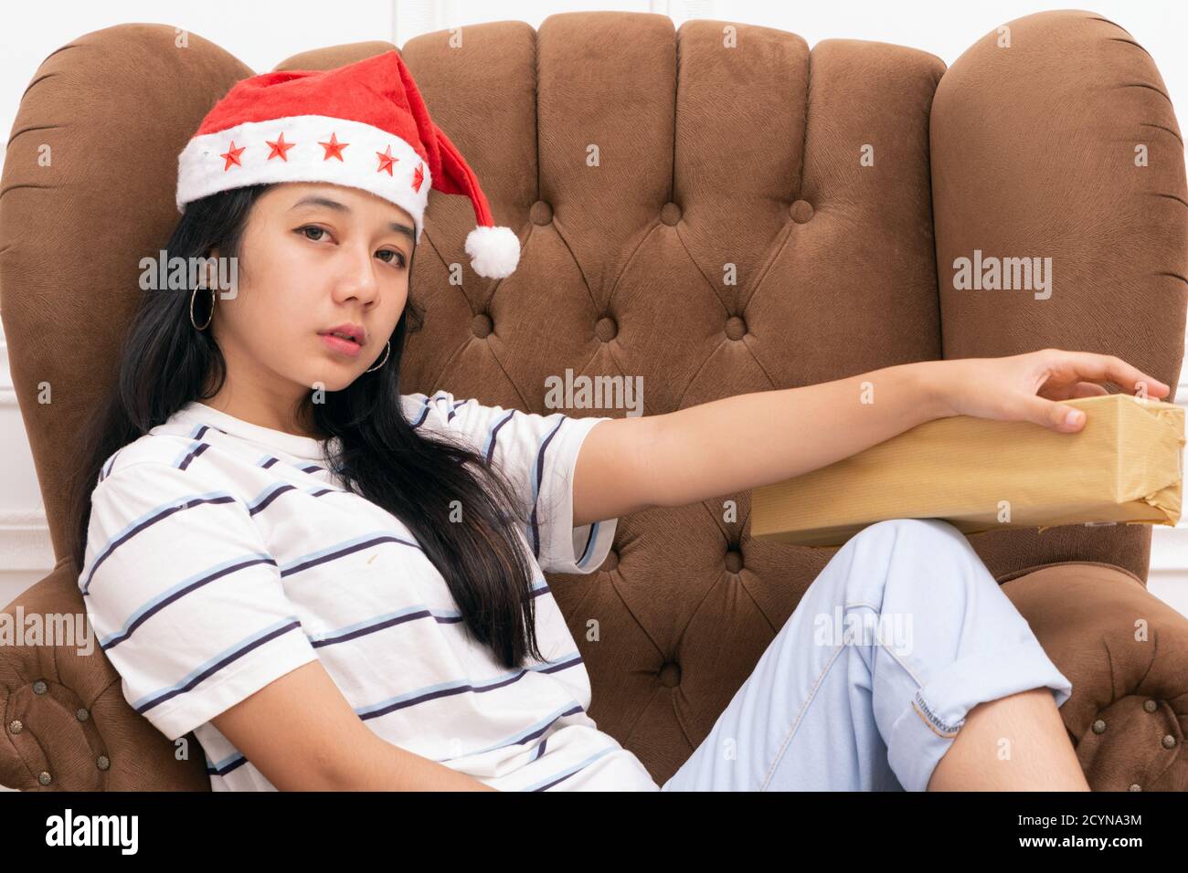 Portrait d'une jeune femme asiatique portant un chapeau de père Noël et de porter des vêtements décontractés assis sur un canapé marron pendant Tenue d'un cadeau avec une décoration de Noël Banque D'Images