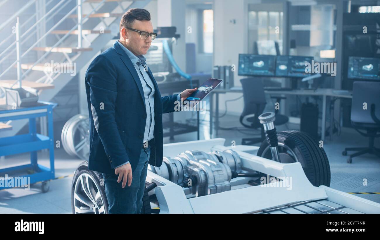 Ingénieur avec lunettes travaille sur une tablette à côté d'un châssis de voiture électrique prototype avec roues, batteries et moteur dans une haute technologie Banque D'Images
