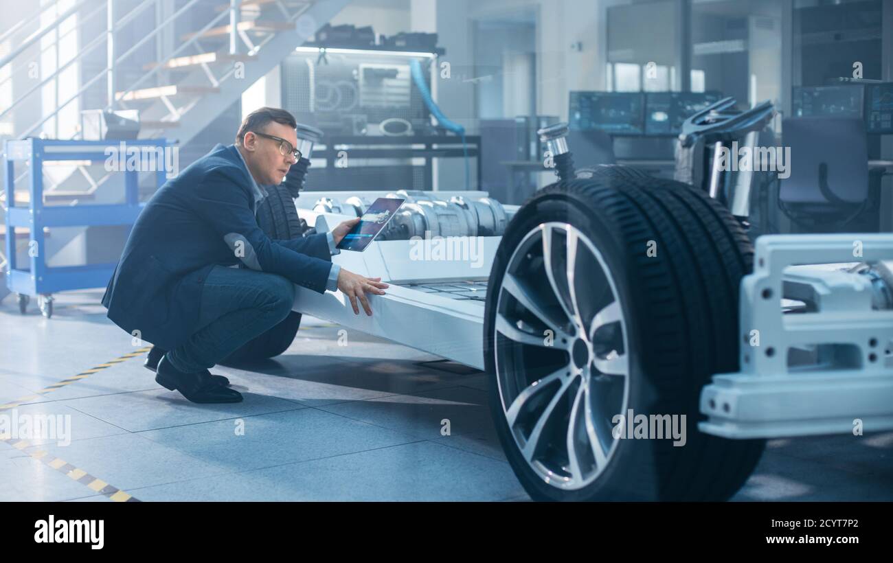 Ingénieur avec lunettes travaille sur une tablette à côté d'un châssis de voiture électrique prototype avec roues, batteries et moteur dans une haute technologie Banque D'Images