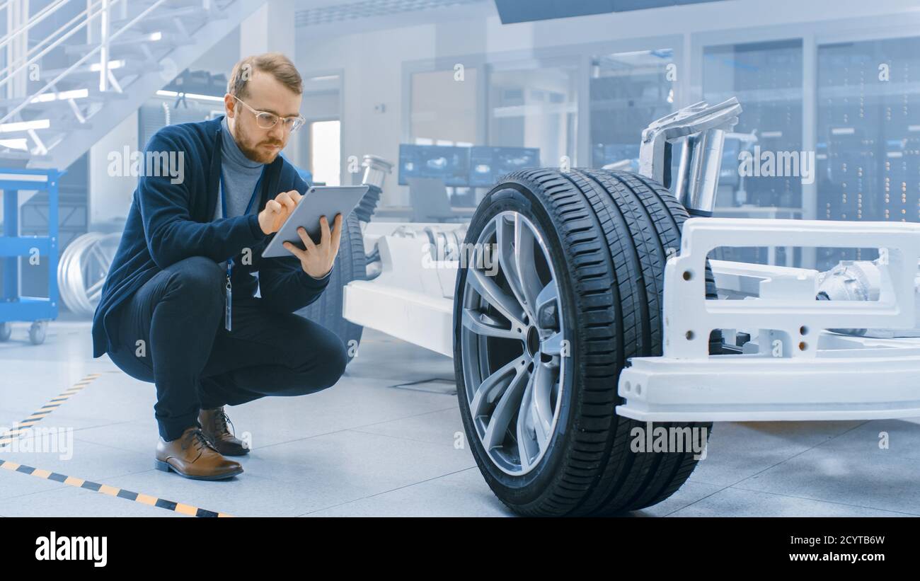 Ingénieur avec des lunettes et Beard travaille sur une tablette ordinateur à côté d'un châssis de voiture électrique prototype avec roues, batteries et moteur dans une haute technologie Banque D'Images
