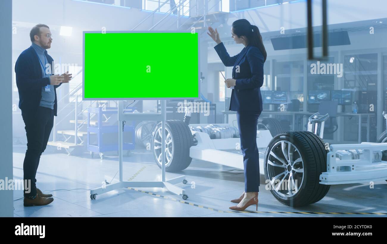 Les ingénieurs concepteurs hommes et femmes discutent de quelque chose sur un tableau blanc interactif avec écran vert à côté d'un prototype de châssis de voiture électrique. Po Banque D'Images