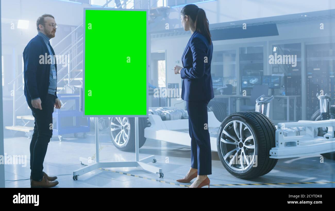 Les ingénieurs concepteurs hommes et femmes discutent de quelque chose sur un tableau blanc interactif avec écran vert à côté d'un prototype de châssis de voiture électrique. Po Banque D'Images