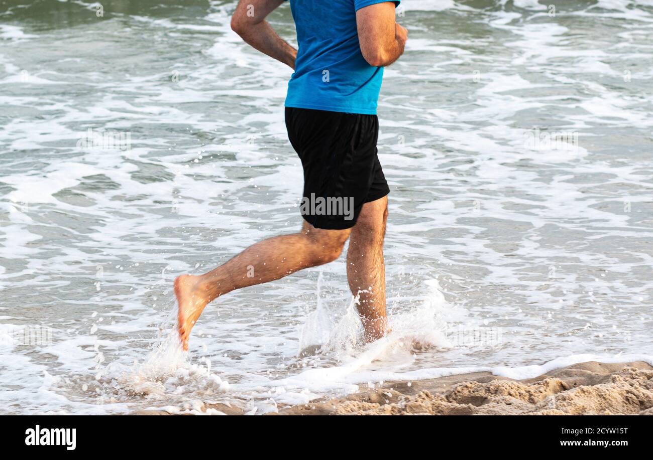 Vue latérale d'un homme courant et éclaboussant dans l'eau au bord de la plage pieds nus. Banque D'Images