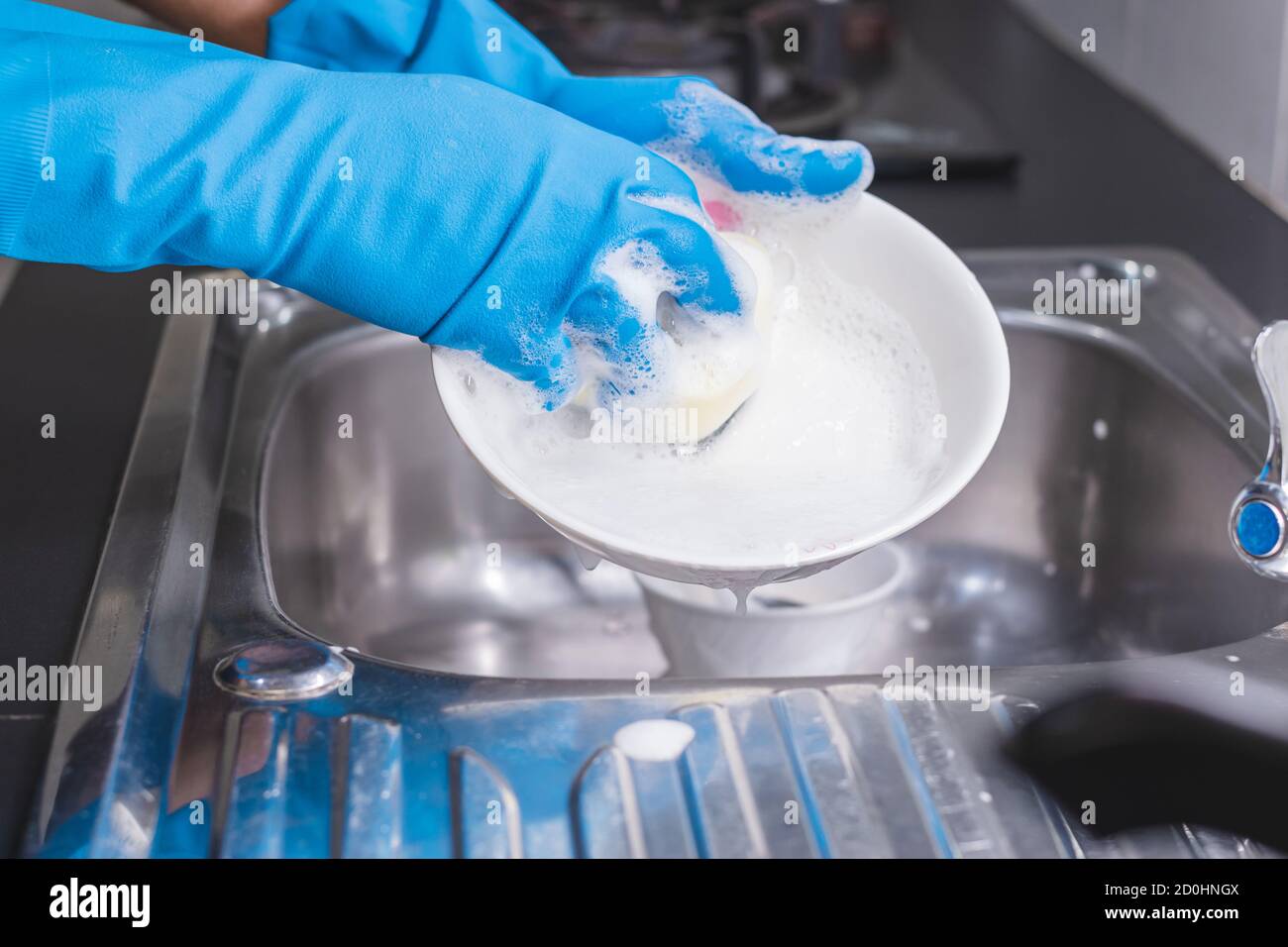 Gros plan d'un homme portant un gant en caoutchouc bleu lavant la tasse avec un liquide vaisselle dans l'évier de la cuisine. Banque D'Images Gros plan d'un homme portant un gant en caoutchouc bleu lavant la tasse avec un liquide vaisselle dans l'évier de la cuisine. Banque D'Images