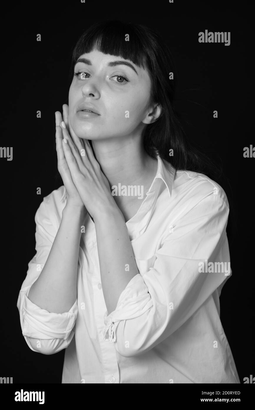 Studio portrait d'une jolie femme brune dans une chemise blanche, sur un fond noir Uni, tenant les deux mains ensemble, près du visage, regardant le Banque D'Images