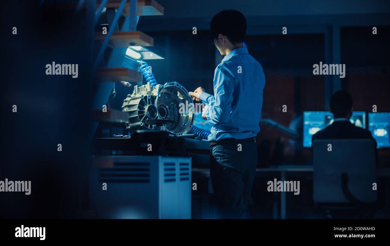L'ingénieur automobile professionnel en lunettes travaille sur les engrenages de transmission dans un laboratoire innovant de haute technologie avec des écrans d'ordinateur. Banque D'Images