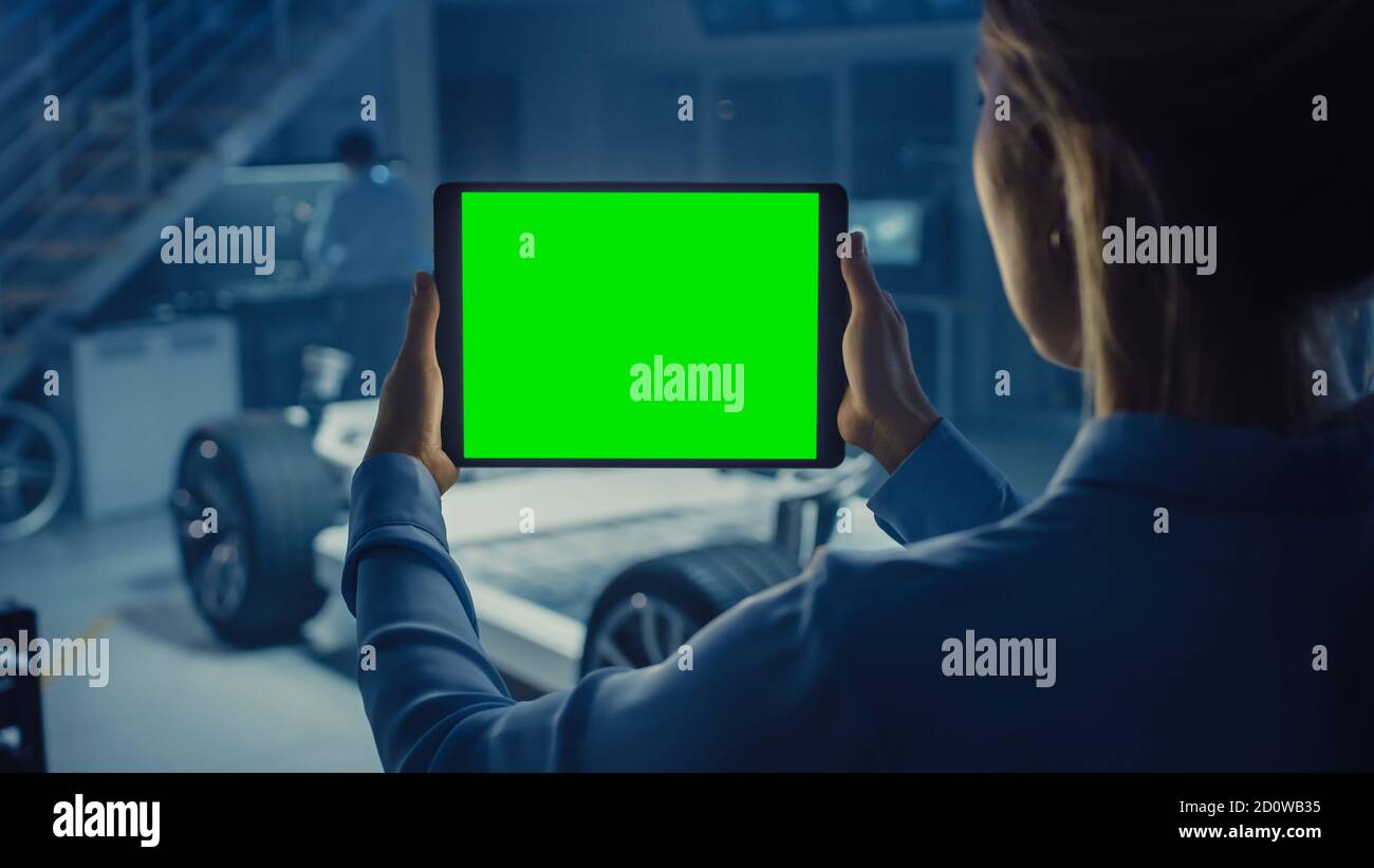 Une femme ingénieur tient un ordinateur tablette avec écran vert Mock Up et regarde le prototype de châssis de voiture électrique avec roues, batteries et moteur Banque D'Images
