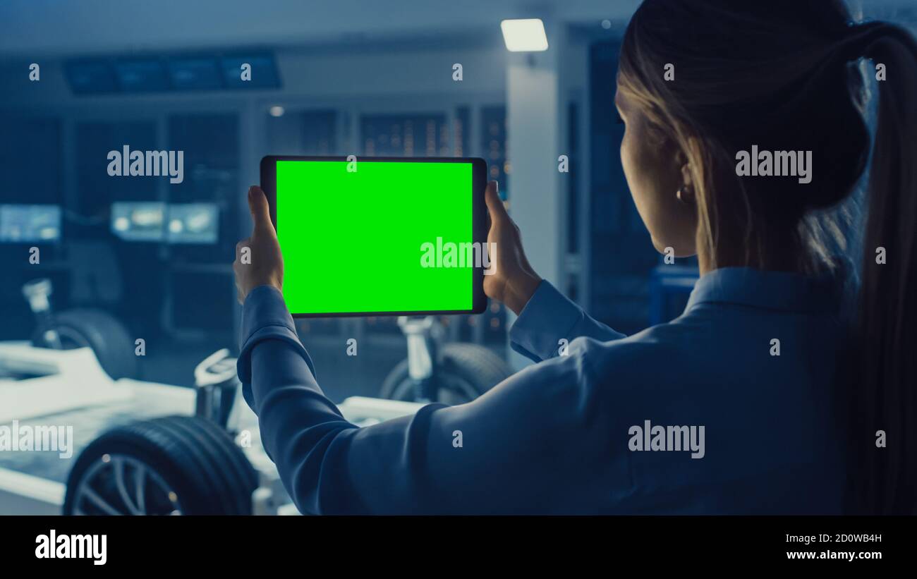 Une femme ingénieur tient un ordinateur tablette avec écran vert Mock Up et regarde le prototype de châssis de voiture électrique avec roues, batteries et moteur Banque D'Images