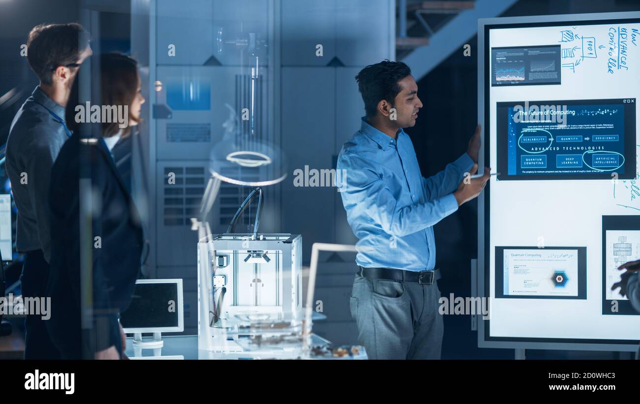 Réunion des ingénieurs dans le laboratoire de recherche technologique : un ingénieur de sexe masculin dirige une présentation à l'aide d'un tableau blanc numérique, montre un modèle de machine, des données Banque D'Images