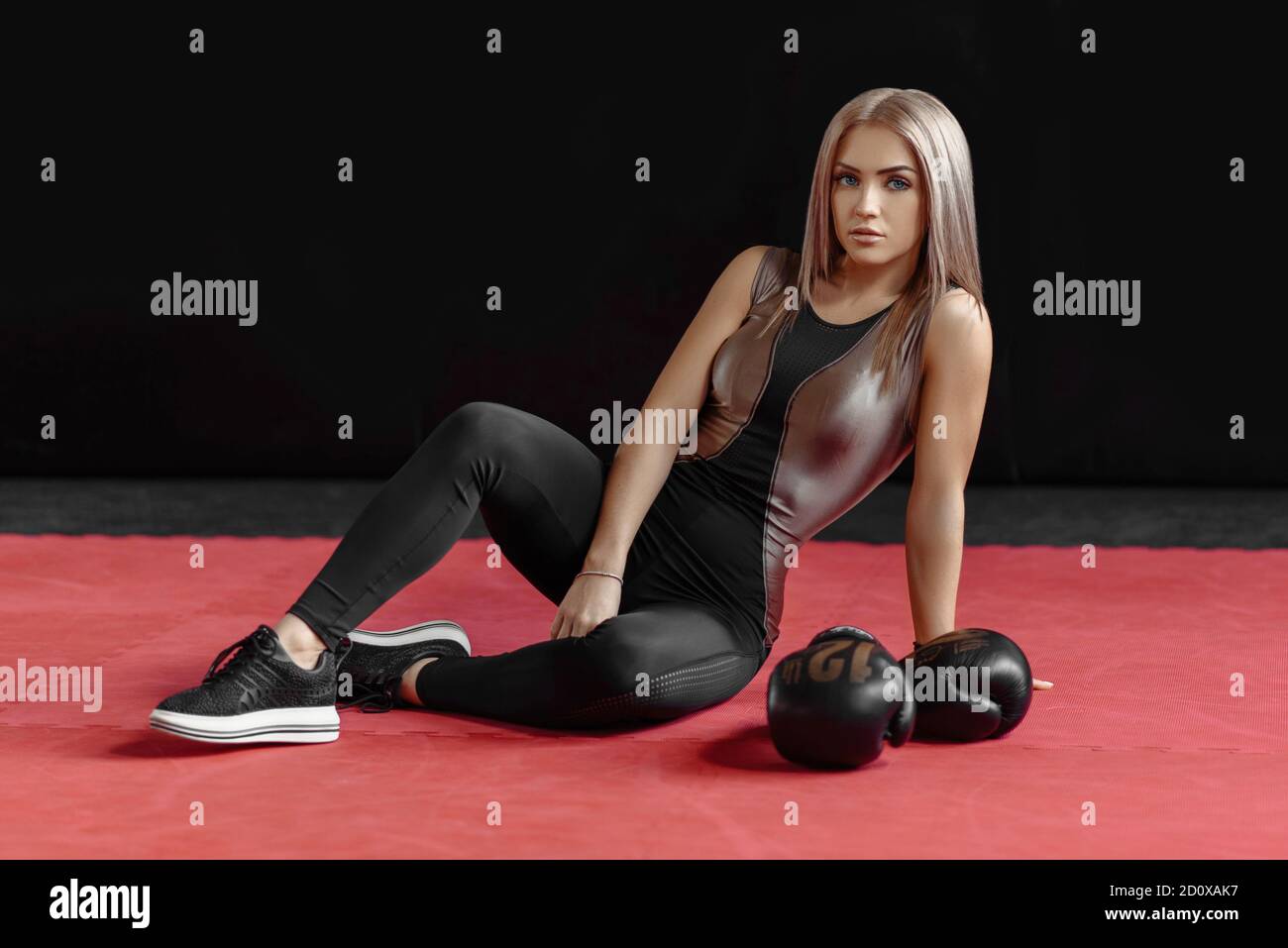 Photo d'une femme en forme physique saine dans des vêtements de sport posant sur un appareil-photo en blanc assis sur le sol avec des gants de boxe noirs pendant l'entraînement dans la salle de gym. Concept de boxe Banque D'Images