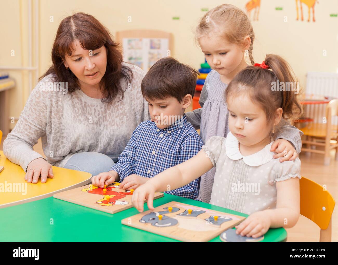 le professeur de l'école primaire recueille le puzzle avec trois élèves assis à la table. Une fille debout derrière Banque D'Images