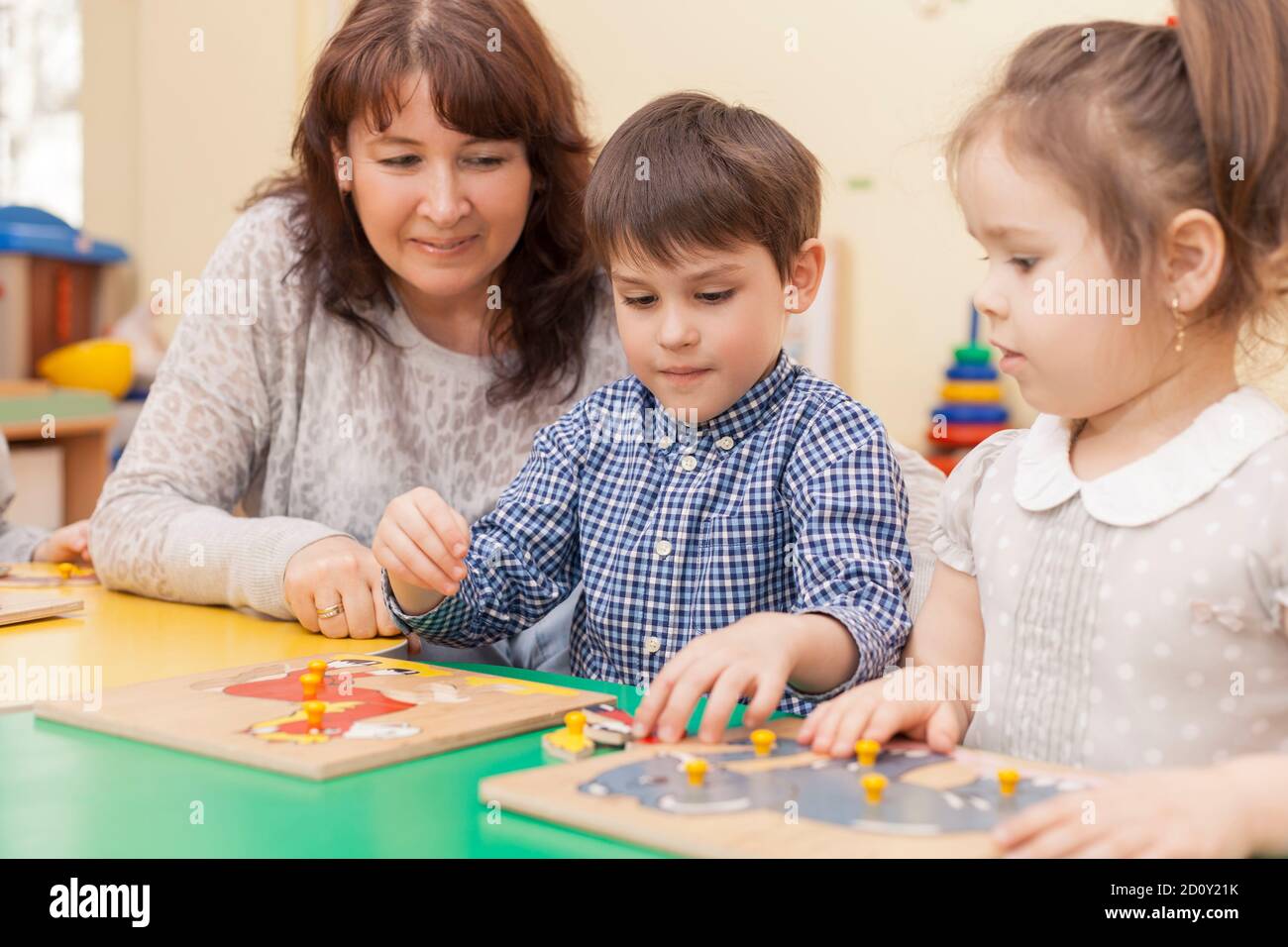 garçon rassemble un puzzle à la table dans la salle de classe, le professeur et la fille le regardant et souriant Banque D'Images