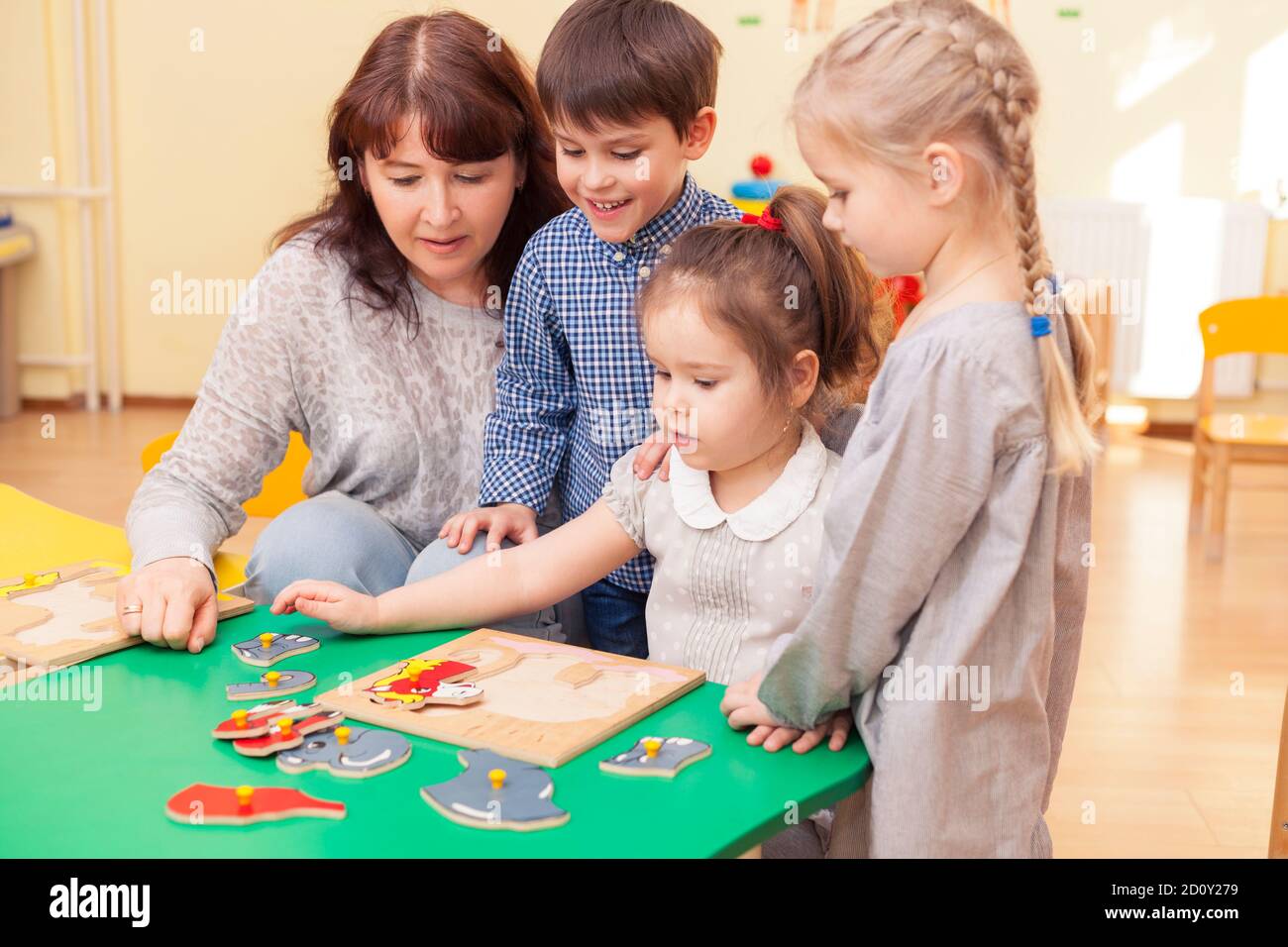 belle femme mature enseignante collectionne le puzzle avec trois élèves d'une école maternelle à la table verte. Image couleur horizontale. Banque D'Images