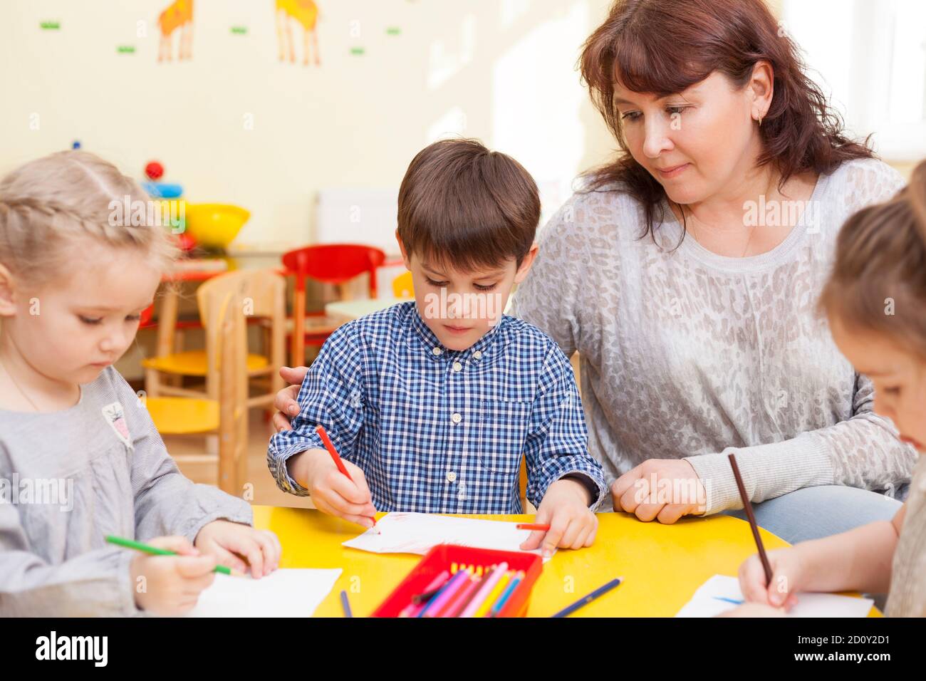 Mignon garçon, élève de la maternelle, dessine à la table jaune, avec les autres élèves. Éducateur, belle femme mûre l'observant et smili Banque D'Images