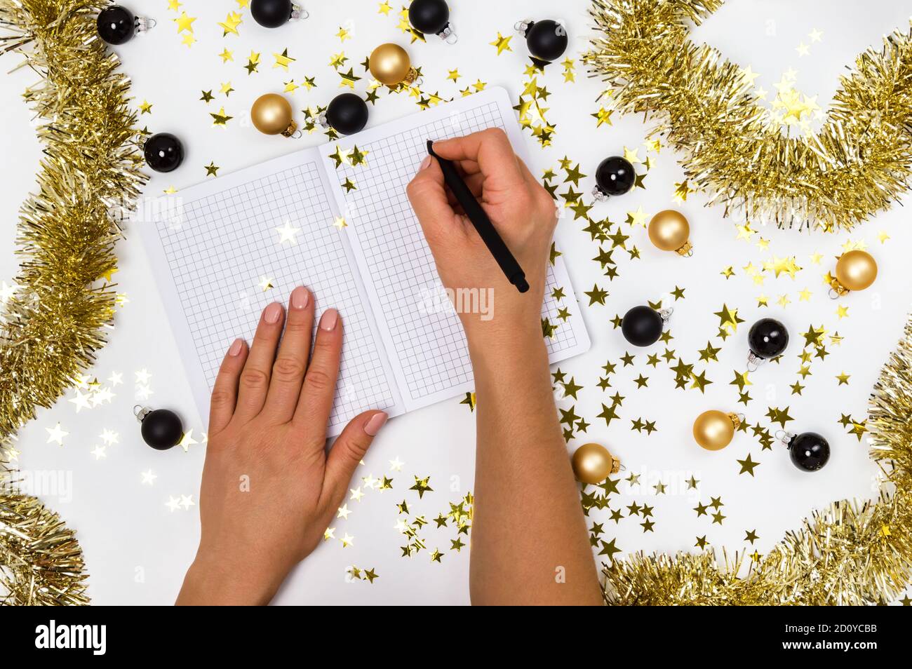 Mains de femmes avec stylo et carnet parmi des boules de Noël dorées et noires, guirlande et confettis étoiles. Liste de souhaits, résolutions du nouvel an ou plan 2021 Banque D'Images