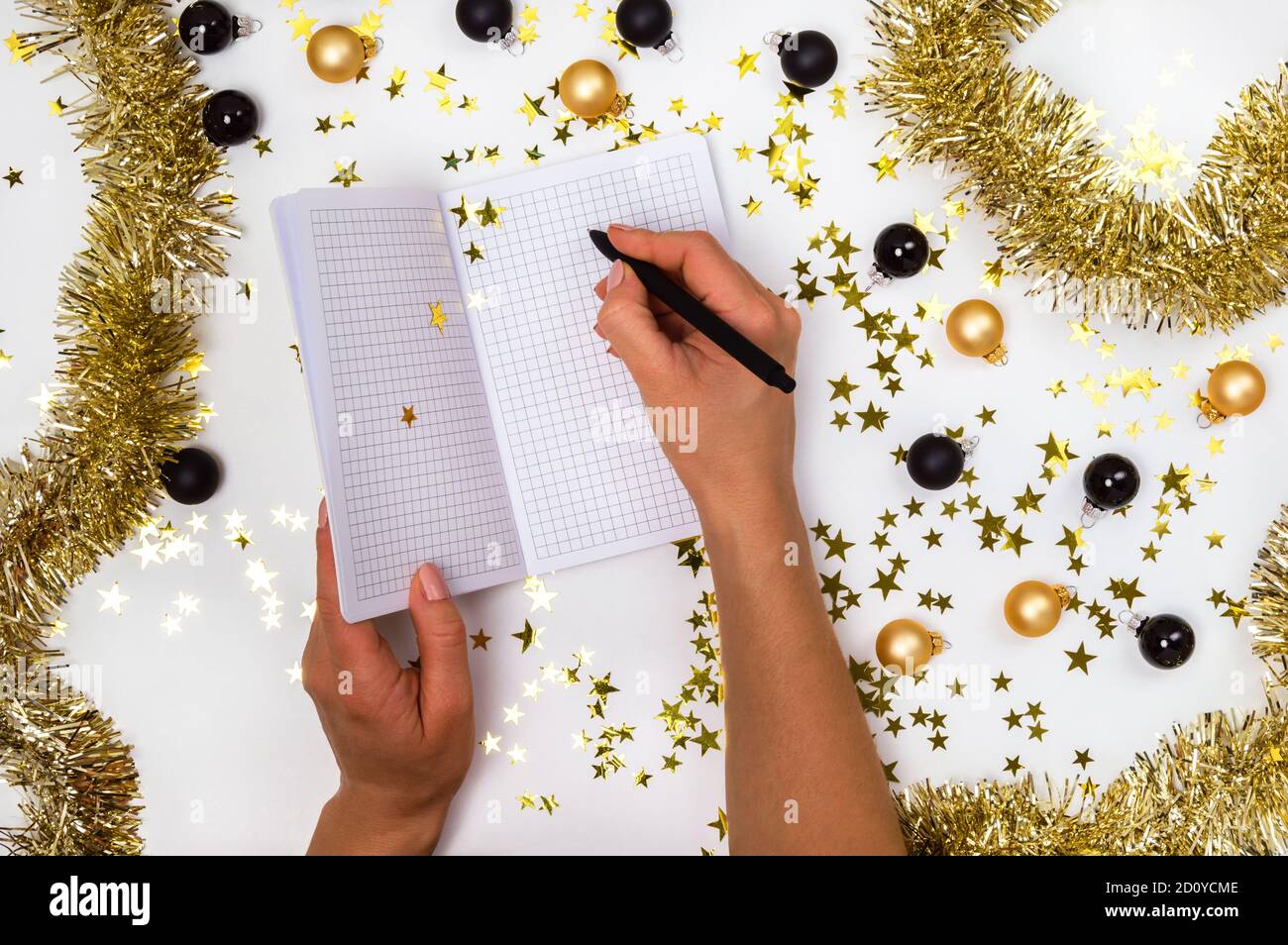Mains de femmes avec stylo et carnet parmi des boules de Noël dorées et noires, guirlande et confettis étoiles. Liste de souhaits, résolutions du nouvel an ou plan 2021 Banque D'Images