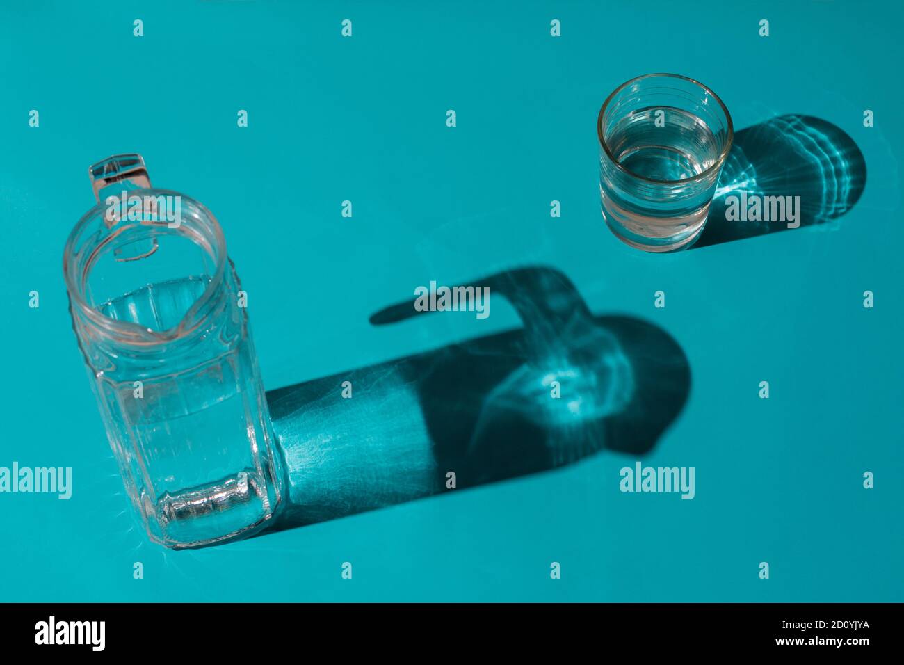 Un verre avec de l'eau à côté d'une verseuse sur un arrière-plan bleu clair où ses ombres sont projetées Banque D'Images