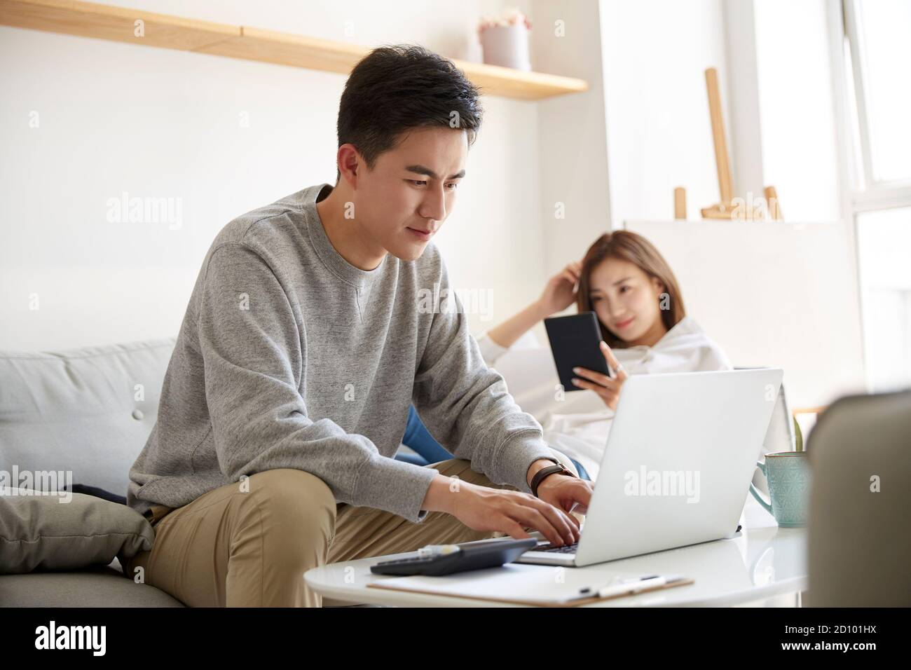 jeune couple asiatique séjournant à la maison avec un mari travaillant avec ordinateur portable et livre de lecture de femme Banque D'Images