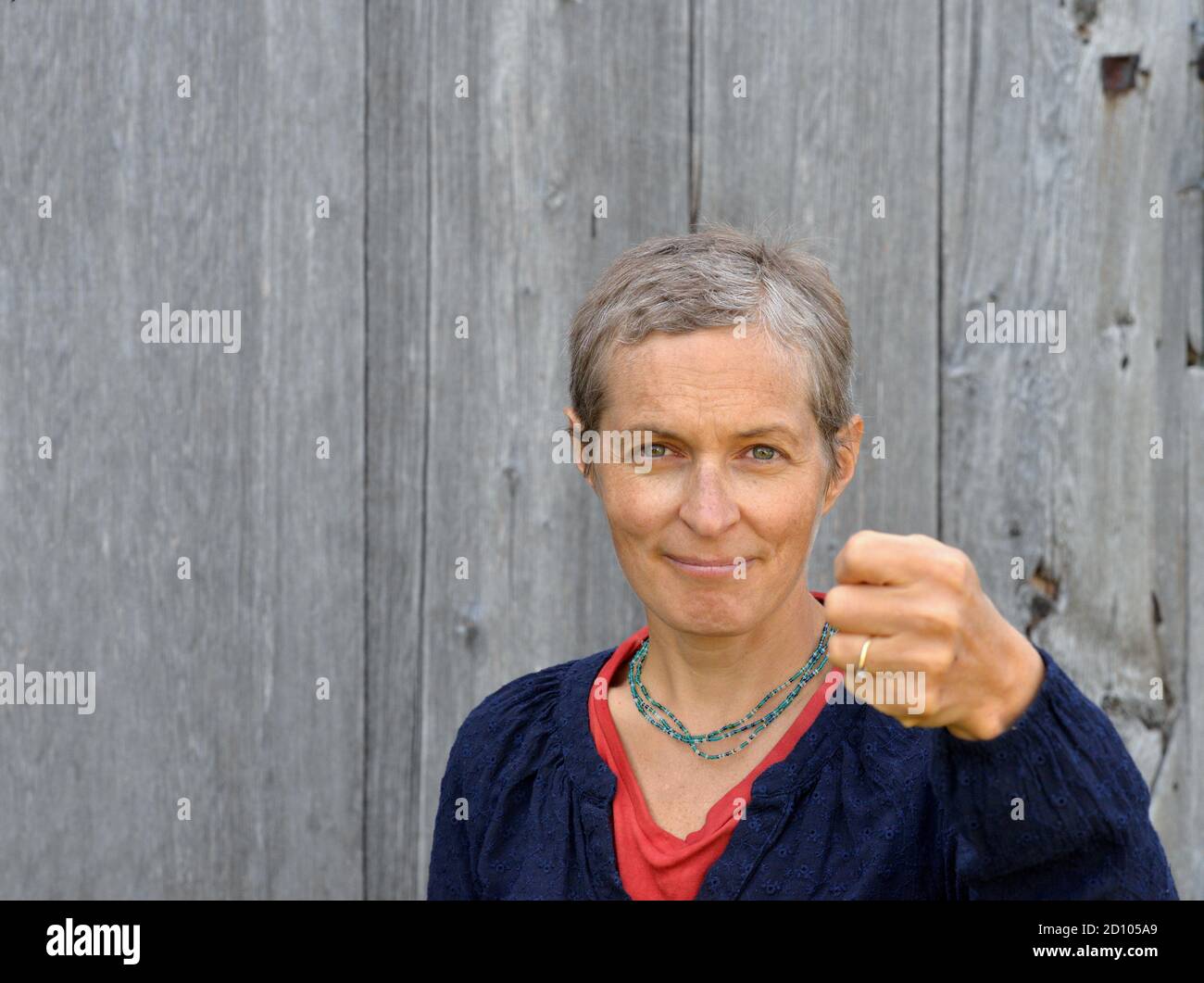 Une femme de campagne caucasienne d'âge moyen sérieuse aux cheveux courts menace le spectateur avec son poing gauche fermé, devant un vieux fond de bois de grange. Banque D'Images