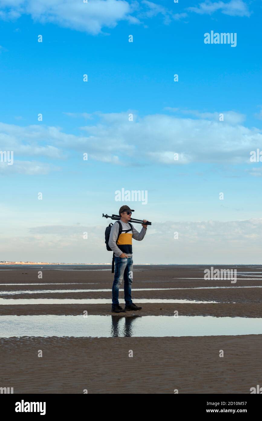 Homme portant un trépied debout sur le sable au bord de la côte. Photographe avec équipement. Banque D'Images