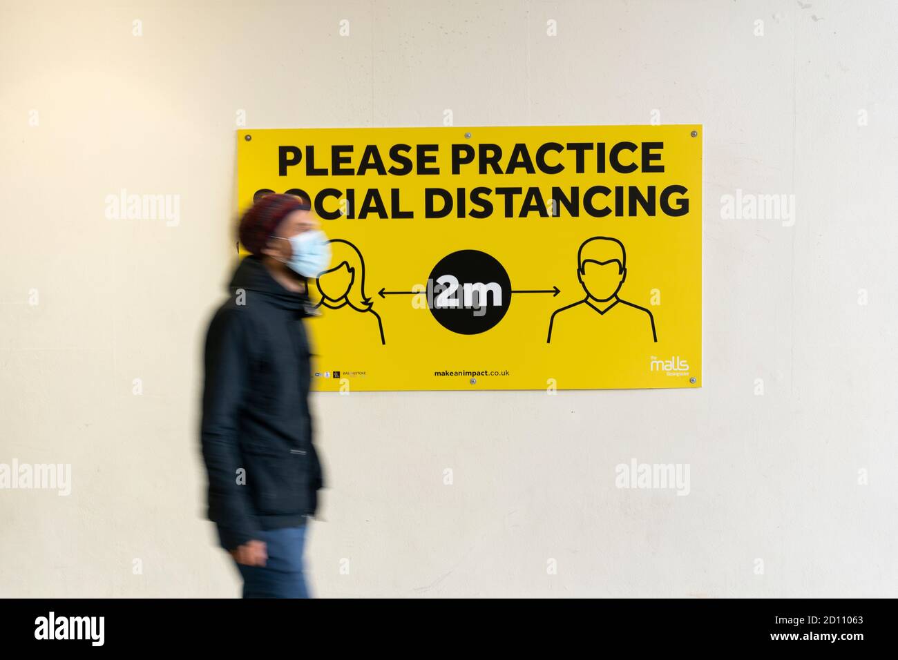 Une personne portant un masque facial marchant devant un panneau disant pratiquer la distanciation sociale pendant la pandémie du coronavirus Covid-19, Basingstoke, Royaume-Uni Banque D'Images
