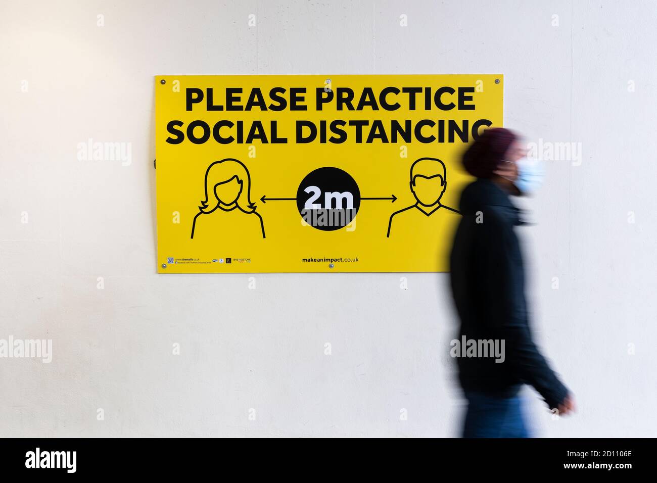 Une personne portant un masque facial marchant devant un panneau disant pratiquer la distanciation sociale pendant la pandémie du coronavirus Covid-19, Basingstoke, Royaume-Uni Banque D'Images